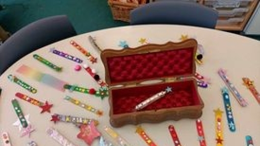 Kidney-shaped table in a classroom covered with decorated wooden sticks and star-shaped ornaments. At the center is an open wooden box with a red velvet interior holding one decorated stick. Chairs and shelves are visible in the background