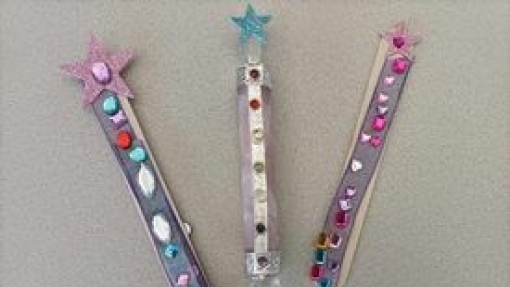 Three handcrafted wands on a light gray surface, each topped with a glittery star and decorated with colorful gems and beads. The left and right wands have pink stars, while the middle has a blue star.