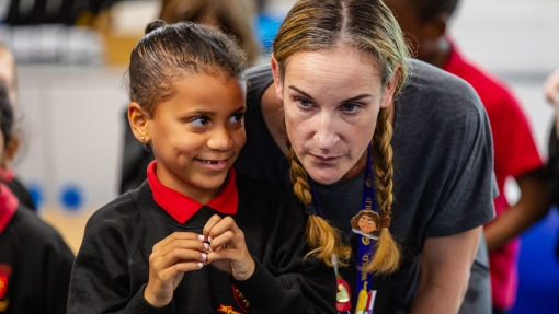 A young student wearing a black school uniform with a red collar is standing next to an adult with braided hair, who is leaning in close. Both of their faces are blurred for privacy. The background shows other students and classroom elements out of focus