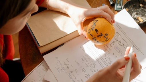 Person holding an orange with the equation 'y = x + 2' written on it, and a white marker in the other hand. On the table are handwritten notes, printed math problems, and a closed book, suggesting a study session focused on functions and derivatives.
