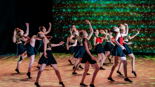 A group of dancers performing on stage in coordinated outfits—black skirts, black socks, and glittery tops in various colors—set against a vibrant, sparkling background. The dancers are mid-movement with arms and legs extended in dynamic poses