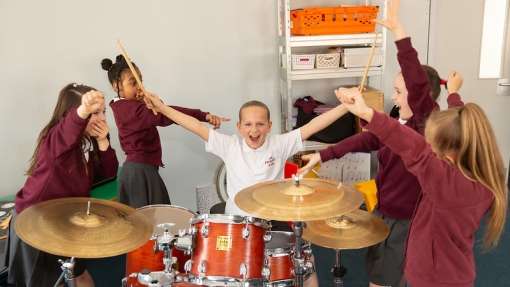 A young person sitting with their arms out wide in front of a drumkit. There are other children around her looking and pointing at her. 