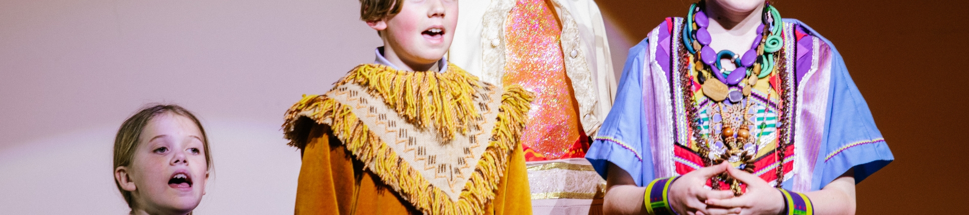 Creative cross-curricular learning at River Beach Primary School. A group of 4 children stood on a stage, wearing colourful costumes with a spotlight pointing at them.