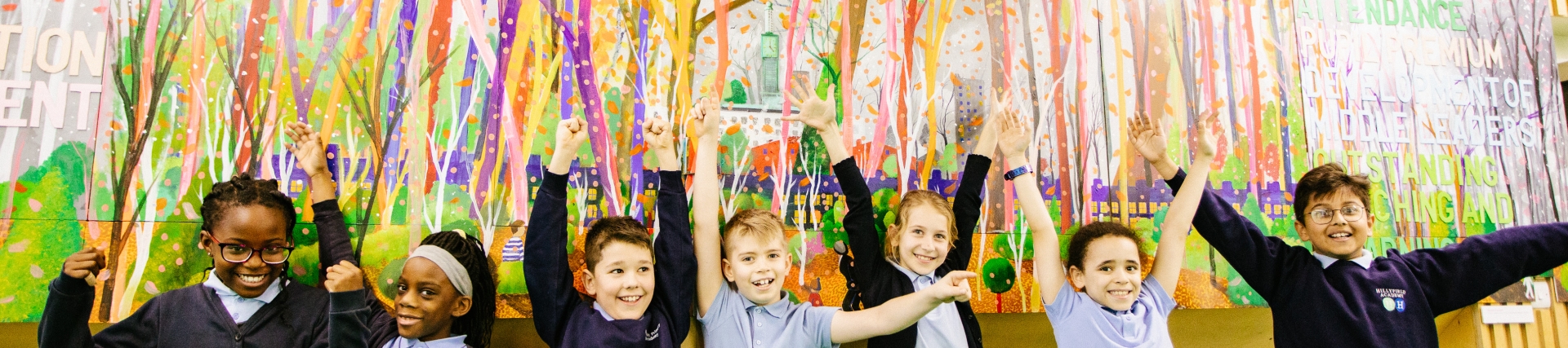 A group of children lined up in front of a large painting on the wall. They are smiling and throwing their arms up in the air.
