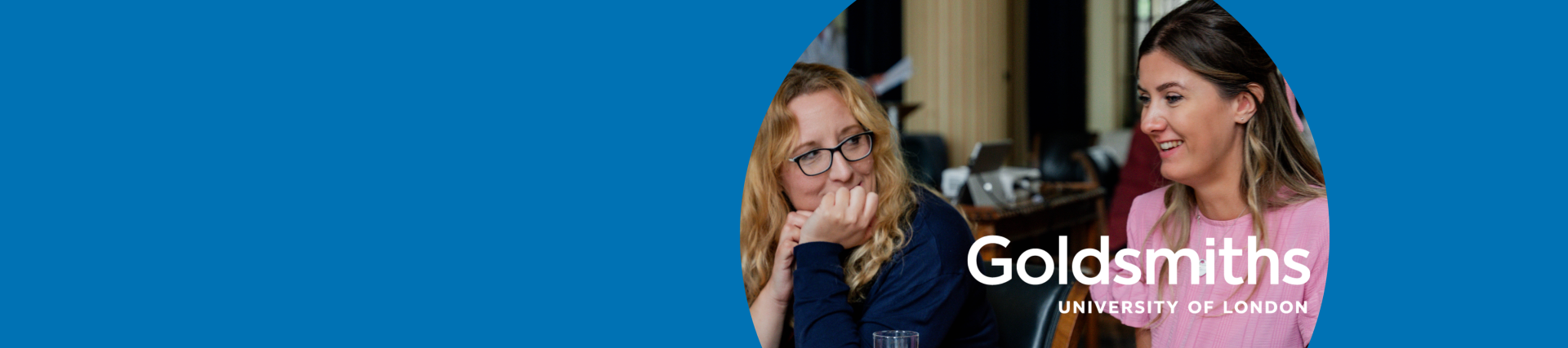 Two white females sitting at a table with documents and post-it notes having a discussion. One lady is looking thoughtful with her hand on her chin. 