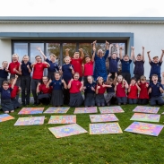 A group of children with an arts practitioner raising hands with joy, their art work on the ground in front of them