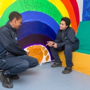Two students sitting in front of a rainbow painted on a wall that says equality 