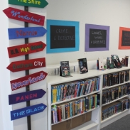 A display of a signpost with different coloured signs pointing in different directions, with books shelves on the right 