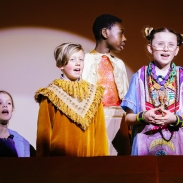 Creative cross-curricular learning at River Beach Primary School. A group of 4 children stood on a stage, wearing colourful costumes with a spotlight pointing at them. 