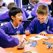 An image of two young people sat on their desk, painting. One child is laughing whilst the other has his head down, concentrating on his painting. 