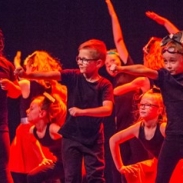 A group of children dance on a stage in dark red lighting. 