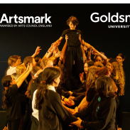 A group of students on a dark stage holding up another student, who is standing on their arms. Artsmark's logo is top left, Goldsmiths logo is top right