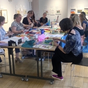 An room with art on the walls and a table and chairs in the middle where a group of women are sitting and taking part in a monoprinting workshop. Lots of art materials are scattered across the table