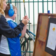 A boy is painting on a large wooden canvas with paper on it. A teacher is standing behind, helping him.