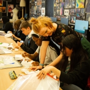 Pupils sat at a long desk and a teacher is leaning over one of the pupils, pointing at their work.