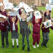 A group of children outside, jumping on the grass, holding up their art work.