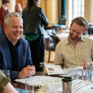 Two men sitting at a table in a meeting room, one is looking down at a document, the other is looking up, smiling.
