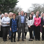 A group of people standing outdoors on a paved area, with trees and a brick building in the background. The group includes men and women in a mix of casual and business attire, some wearing name tags or lanyards.