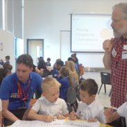 A classroom with children seated at tables, working on large sheets of paper using markers. Two adults—one in a blue shirt and one in a red checkered shirt—are assisting them. A projector screen at the front displays the text 'Young Creativity Champions.' Natural light enters through tall windows on the left.