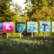 Six colorful signs on a grassy field spell out 'CREATE,' each letter painted uniquely on square canvases mounted on wooden stakes, with lush green trees in the background.