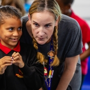 A young student wearing a black school uniform with a red collar is standing next to an adult with braided hair, who is leaning in close. Both of their faces are blurred for privacy. The background shows other students and classroom elements out of focus