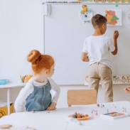 Three children in a classroom: one stands at a whiteboard drawing or writing, while two others sit at a table working with small objects and papers. The whiteboard displays colorful drawings and numbers from 5 to 10.