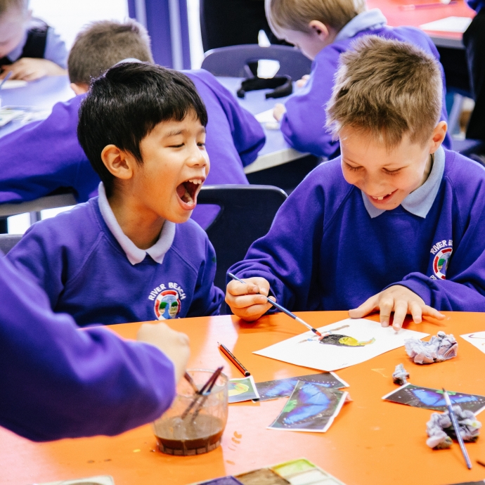 An image of two young people sat on their desk, painting. One child is laughing whilst the other has his head down, concentrating on his painting. 